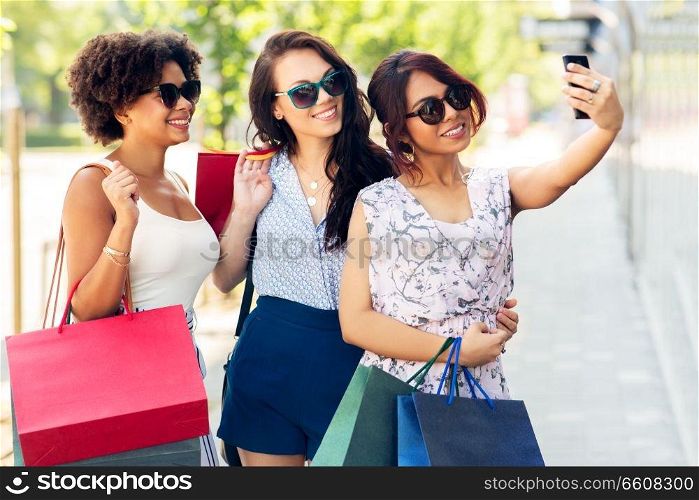 sale, consumerism and technology concept - happy young women with shopping bags taking selfie by smartphone in city. women with shopping bags taking selfie in city