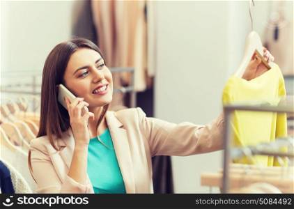 sale, consumerism, fashion, communication and people concept - happy young woman with shopping bags choosing clothes and calling on smartphone in mall or clothing store. woman calling on smartphone at clothing store. woman calling on smartphone at clothing store