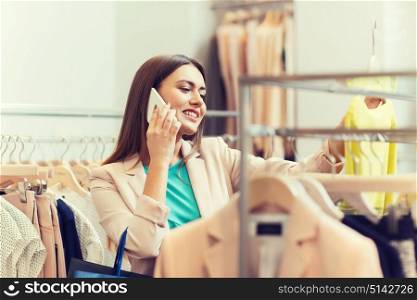 sale, consumerism, fashion, communication and people concept - happy young woman with shopping bags choosing clothes and calling on smartphone in mall or clothing store. woman calling on smartphone at clothing store