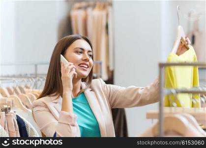 sale, consumerism, fashion, communication and people concept - happy young woman with shopping bags choosing clothes and calling on smartphone in mall or clothing store