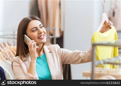 sale, consumerism, fashion, communication and people concept - happy young woman with shopping bags choosing clothes and calling on smartphone in mall or clothing store