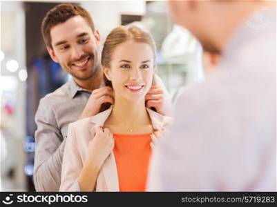 sale, consumerism, presents, holidays and people concept - happy couple trying golden pendant on at jewelry store in mall