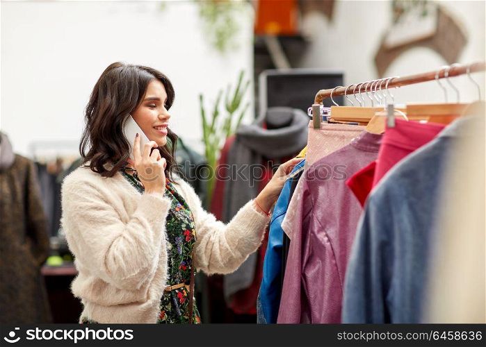 sale, consumerism, shopping and people concept - happy young woman choosing clothes and calling on smartphone at vintage clothing store. woman calling on smartphone at clothing store