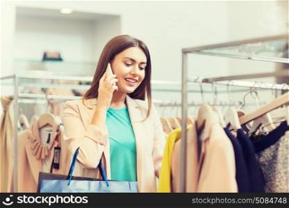 sale, consumerism, shopping and people concept - happy young woman with shopping bags choosing clothes and calling on smartphone in mall or clothing store. happy young woman choosing clothes in mall. happy young woman choosing clothes in mall