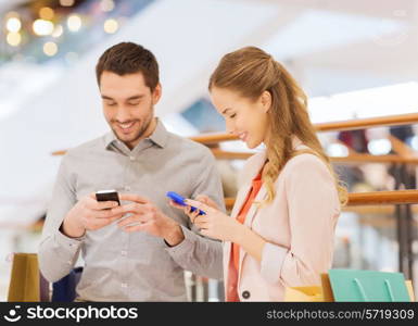 sale, consumerism, technology and people concept - happy young couple with shopping bags and smartphones in mall