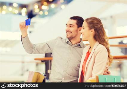 sale, consumerism, technology and people concept - happy young couple with shopping bags and smartphone taking selfie in mall