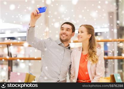 sale, consumerism, technology and people concept - happy young couple with shopping bags and smartphone taking selfie in mall with snow effect
