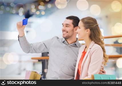 sale, consumerism, technology and people concept - happy young couple with shopping bags and smartphone taking selfie in mall. happy couple with smartphone taking selfie in mall