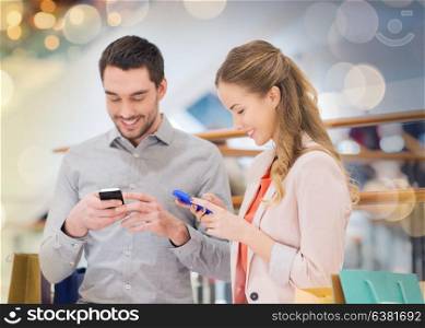 sale, consumerism, technology and people concept - happy young couple with shopping bags and smartphones in mall. couple with smartphones and shopping bags in mall