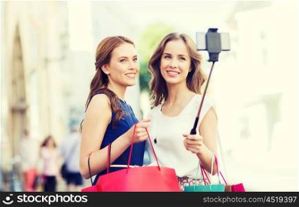sale, consumerism, technology and people concept - happy young women with shopping bags and smartphone selfie stick taking picture on city street