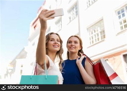 sale, consumerism, technology and people concept - happy young women with shopping bags and smartphone taking selfie on city street. women shopping and taking selfie by smartphone. women shopping and taking selfie by smartphone
