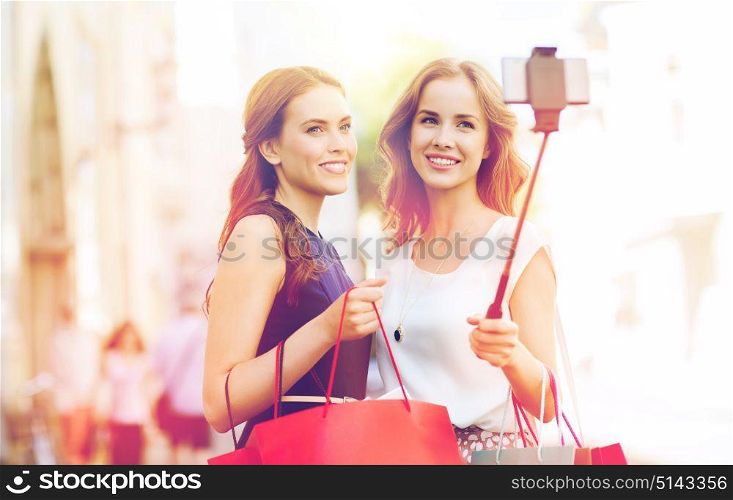 sale, consumerism, technology and people concept - happy young women with shopping bags and smartphone selfie stick taking picture on city street. happy women with shopping bags and smartphone
