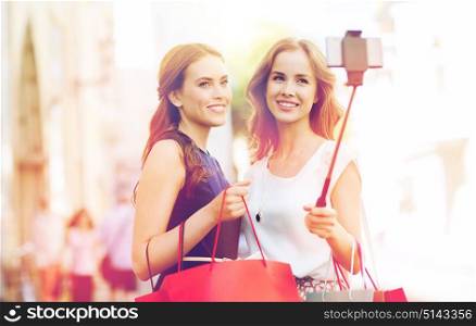 sale, consumerism, technology and people concept - happy young women with shopping bags and smartphone selfie stick taking picture on city street. happy women with shopping bags and smartphone