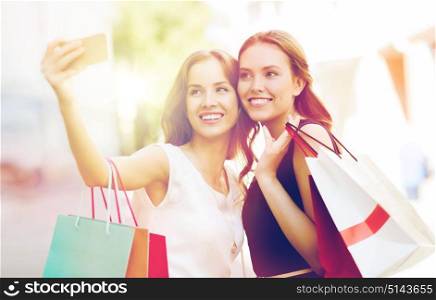 sale, consumerism, technology and people concept - happy young women with shopping bags and smartphone taking selfie on city street. happy women with shopping bags and smartphone