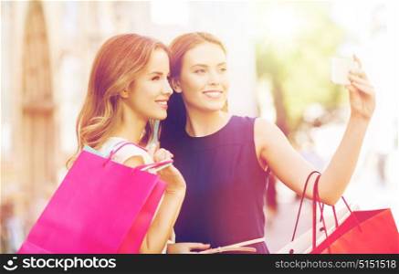 sale, consumerism, technology and people concept - happy young women with shopping bags and smartphone taking selfie on city street. happy women with shopping bags and smartphone