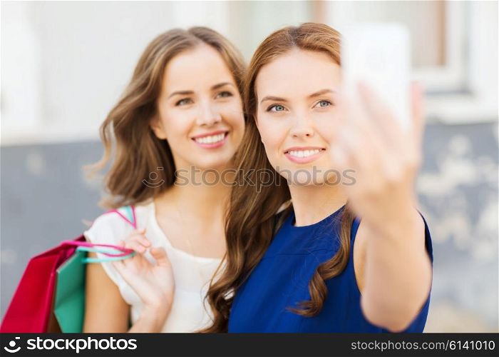sale, consumerism, technology and people concept - happy young women with shopping bags and smartphone taking selfie on city street