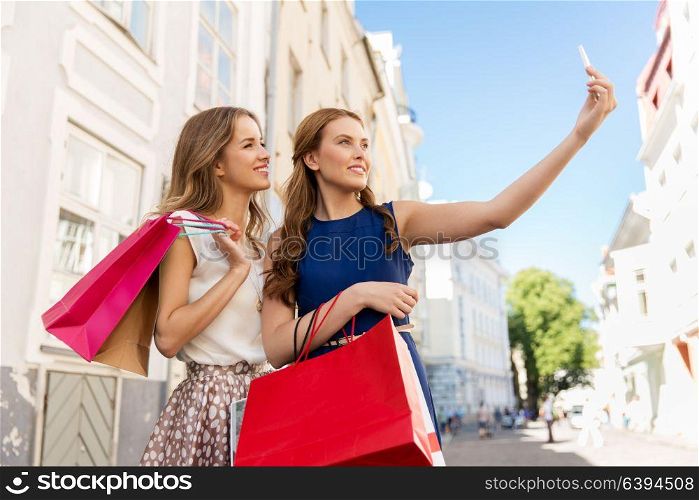 sale, consumerism, technology and people concept - happy young women with shopping bags and smartphone taking selfie on city street. women shopping and taking selfie by smartphone