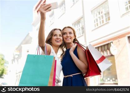 sale, consumerism, technology and people concept - happy young women with shopping bags and smartphone taking selfie on city street. women shopping and taking selfie by smartphone