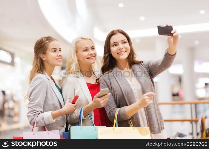 sale, consumerism, technology and people concept - happy young women with smartphones and shopping bags taking selfie in mall