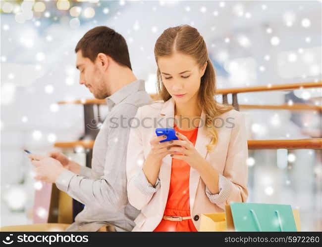 sale, consumerism, technology and people concept - young couple with shopping bags and smartphones texting message in mall with snow effect