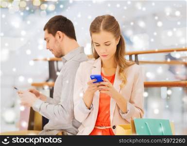 sale, consumerism, technology and people concept - young couple with shopping bags and smartphones texting message in mall with snow effect