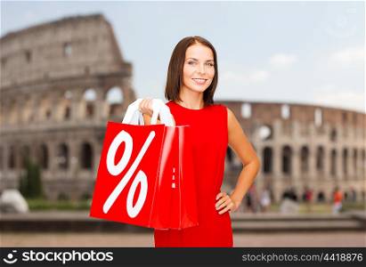 sale, discount, tourism and holidays concept - smiling young woman in red dress with shopping bags with percent sign over coliseum background