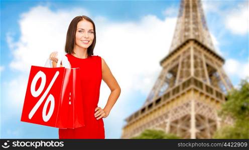 sale, discount, tourism and holidays concept - smiling young woman in red dress with shopping bags with percent sign over paris eiffel tower background