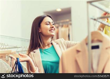 sale, fashion, consumerism and people concept - happy young woman with shopping bags choosing clothes in mall or clothing store. happy young woman choosing clothes in mall