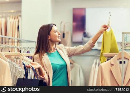 sale, fashion, consumerism and people concept - happy young woman with shopping bags choosing clothes in mall or clothing store. happy young woman choosing clothes in mall