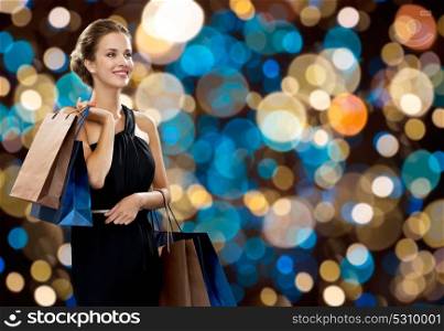 sale, fashion, people and luxury concept - happy beautiful young woman in black dress with shopping bags over holidays lights background. happy woman in black dress with shopping bags