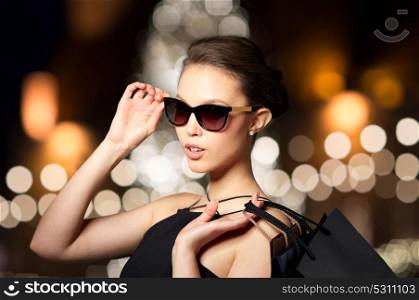 sale, fashion, people and luxury concept - happy beautiful young woman in black sunglasses with shopping bags over christmas tree lights background. woman in shades with shopping bags at christmas