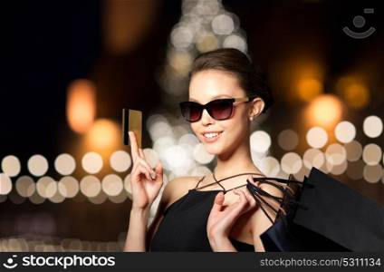 sale, fashion, people and luxury concept - happy beautiful young woman in black sunglasses with credit card and shopping bags over christmas tree lights background. woman with credit card and shopping bags