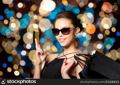 sale, fashion, people and luxury concept - happy beautiful young woman in black sunglasses with credit card and shopping bags over holidays lights background. happy woman with credit card and shopping bags