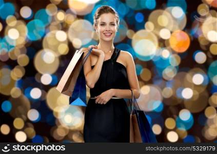 sale, fashion, people and luxury concept - happy beautiful young woman in black dress with shopping bags over holidays lights background. happy woman in black dress with shopping bags