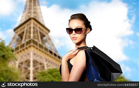 sale, fashion, tour, people and luxury concept - happy beautiful young woman in black sunglasses with shopping bags over paris eiffel tower background