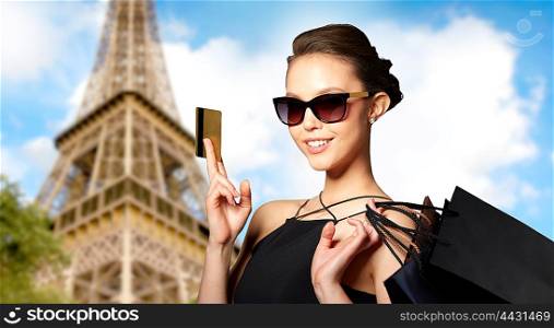 sale, finances, fashion, people and luxury concept - happy beautiful young woman in black sunglasses with credit card and shopping bags over paris eiffel tower background