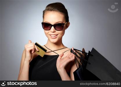 sale, finances, fashion, people and luxury concept - happy beautiful young woman in black sunglasses with credit card and shopping bags over gray background