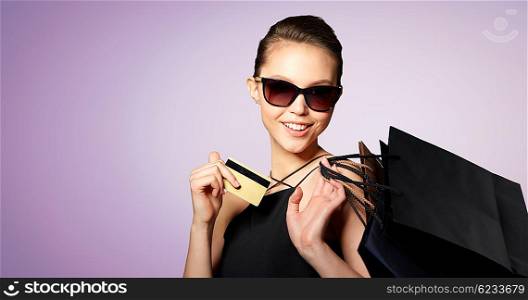 sale, finances, fashion, people and luxury concept - happy beautiful young woman in black sunglasses with credit card and shopping bags over violet background