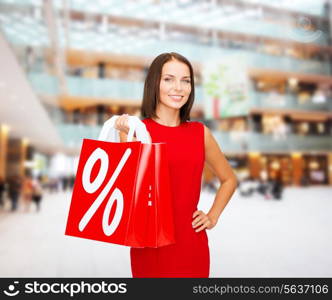 sale, gifts, christmas, holidays and people concept - smiling woman in red dress with shopping bags and percent sign over shopping center background