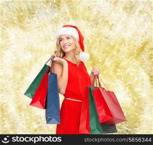 sale, gifts, christmas, holidays and people concept - smiling woman in red dress and santa helper hat with shopping bags over yellow lights background
