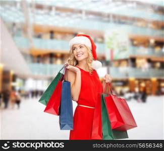 sale, gifts, christmas, holidays and people concept - smiling woman in red dress and santa helper hat with shopping bags over mall background