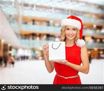 sale, gifts, christmas, holidays and people concept - smiling woman in red dress and santa helper hat with white blank shopping bag over mall background