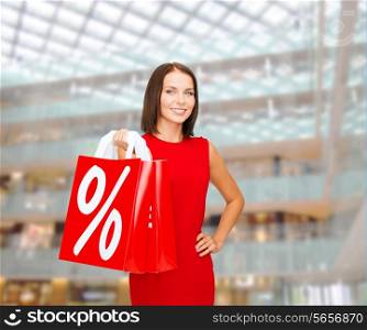 sale, gifts, christmas, holidays and people concept - smiling woman in red dress with shopping bags and percent sign over shopping center background