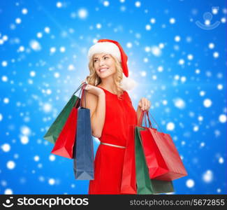 sale, gifts, christmas, holidays and people concept - smiling woman in red dress and santa helper hat with shopping bags over blue snowing background