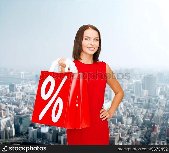 sale, gifts, christmas, holidays and people concept - smiling woman in red dress with shopping bags and percent sign over city background
