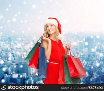 sale, gifts, christmas, holidays and people concept - smiling woman in red dress and santa helper hat with shopping bags over snowy city background