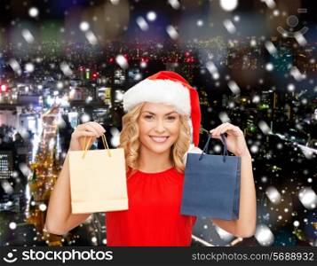 sale, gifts, christmas, holidays and people concept - smiling woman in red dress and santa helper hat with shopping bags over snowy night city background