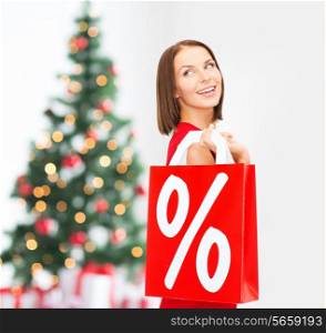 sale, gifts, holidays and people concept - smiling woman in red dress holding shopping bags with percent sign over living room and christmas tree background