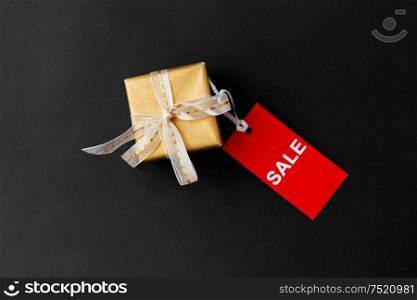 sale, holidays and christmas concept - small golden gift box and red discount tag on black background. small gift box and red sale tag with discount sign