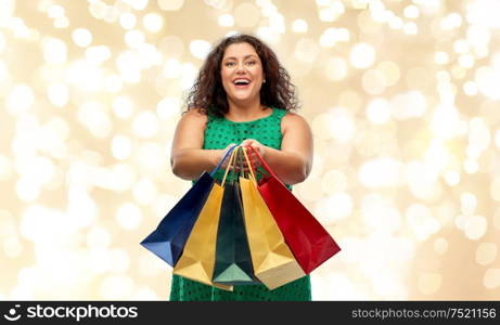 sale, holidays and consumerism concept - happy woman in green dress with shopping bags over festive lights on beige background. happy woman with shopping bags over lights